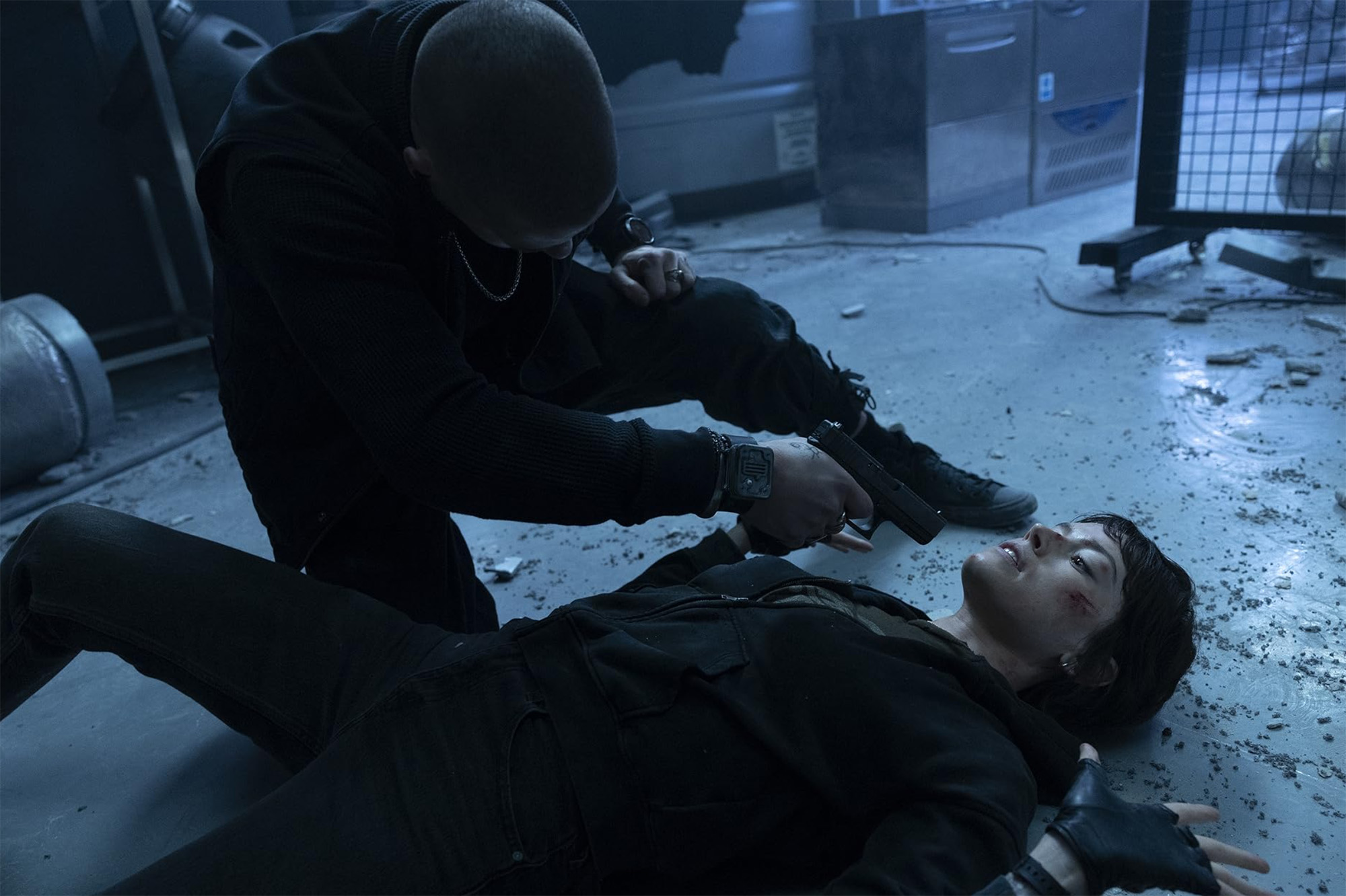 A film still from "Cleaner" showing Daisy Ridley lying injured on the floor in a dim, industrial setting, as Taz Skylar kneels over her, holding a gun to her face.