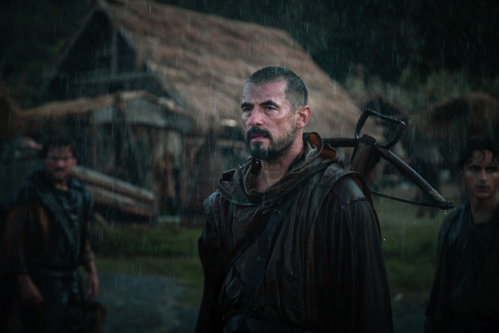 A promotional still for "William Tell" showing Claes Bang in costume as the title character, standing in front of a stormy mountain backdrop with a crossbow slung over his shoulder and a determined expression.