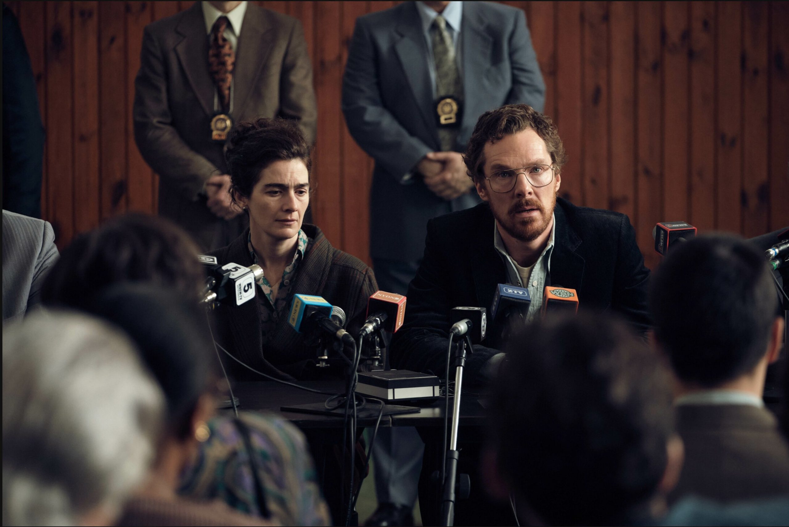 A scene from "Eric" showing Benedict Cumberbatch seated at a press table surrounded by microphones, alongside a woman in formal attire. They address an off-camera audience while uniformed officers stand behind them in front of a wood-panelled wall.