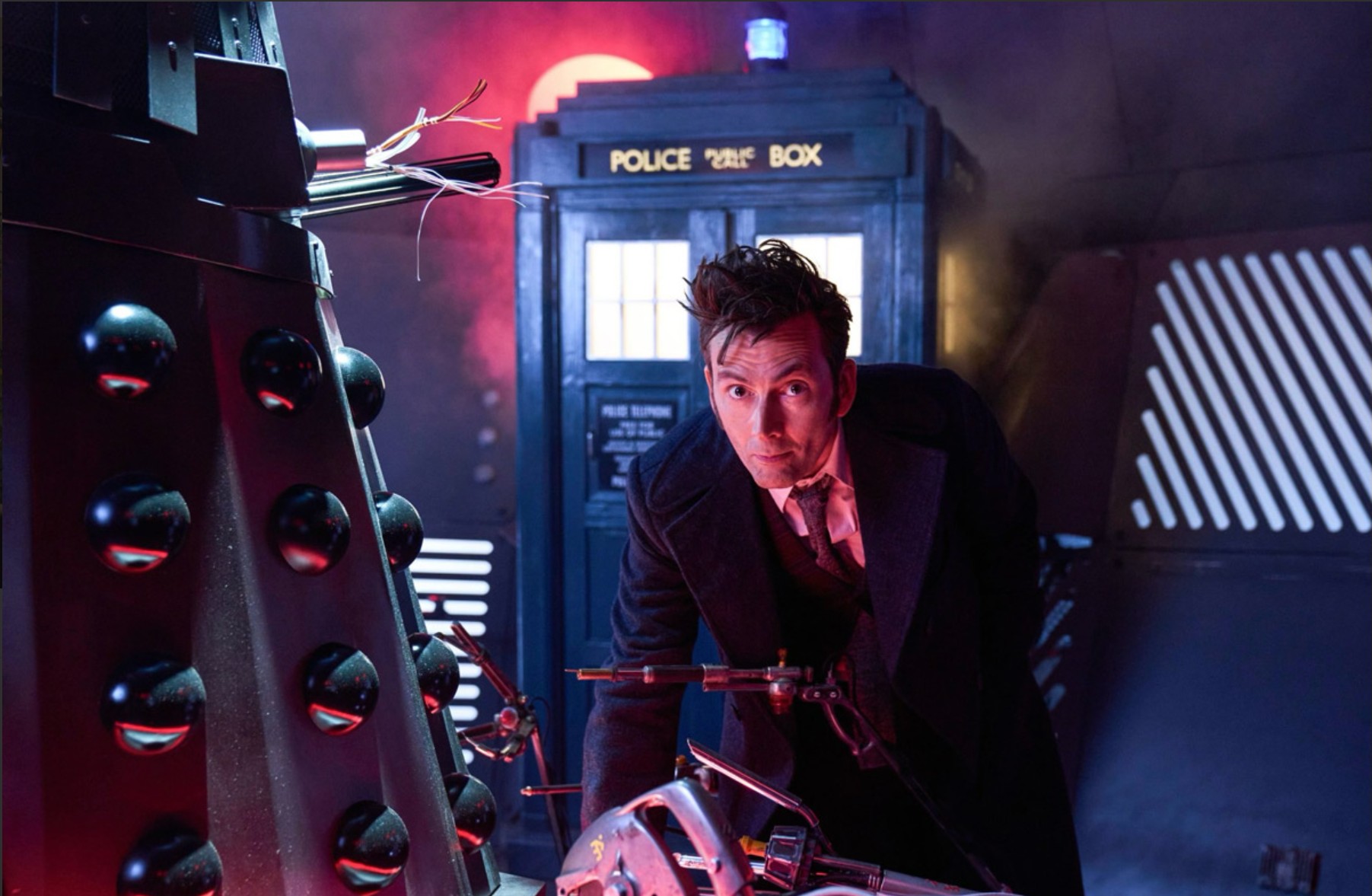 A scene from "Doctor Who" featuring David Tennant as the Doctor inside a futuristic control room, crouched beside a damaged Dalek with the TARDIS illuminated behind him in blue and red lighting.